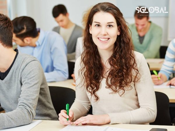 Nữ sinh mỉm cười tự tin trong lớp học với bạn bè quốc tế xung quanh, thể hiện tinh thần lạc quan và sự hòa nhập của du học sinh.