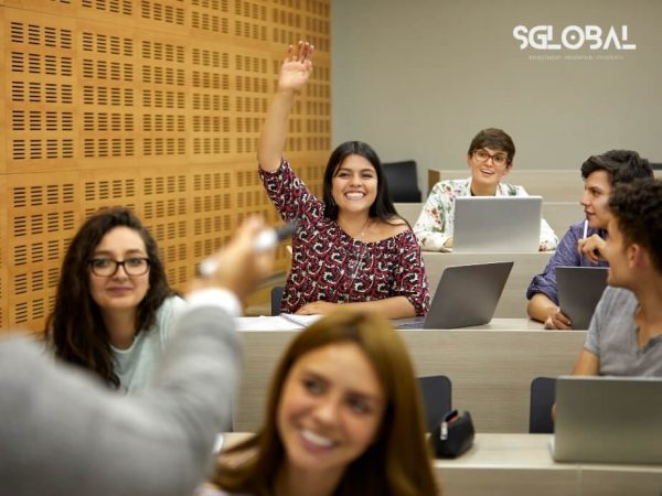 Sinh viên trong lớp học đang giơ tay phát biểu, cười nói sôi nổi với nhau – thể hiện môi trường học tập thân thiện.