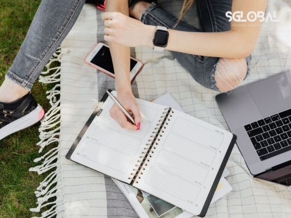 Sinh viên ghi chép vào sổ tay khi học tập ngoài trời, với laptop và sách – biểu tượng học tập linh hoạt tại Mỹ.